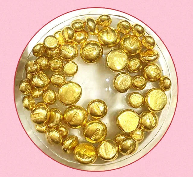 A collection of shiny gold coin-shaped craft embellishments arranged in a circle inside a clear plastic container, set against a solid pink background.