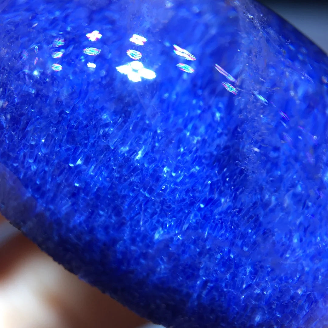 A close-up of a glossy, deep blue gemstone or polished crystal with visible internal inclusions and a textured, fibrous appearance under bright light.