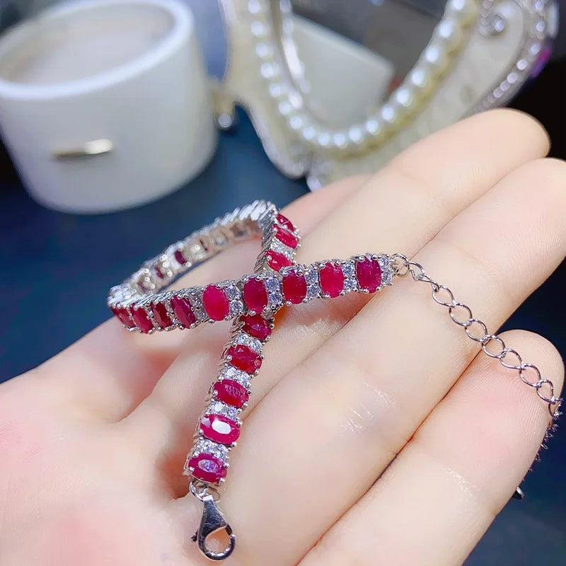 A silver-toned cross pendant bracelet adorned with oval red gemstones and small clear stones, held in a person's hand with a blurred mirror and white container in the background.