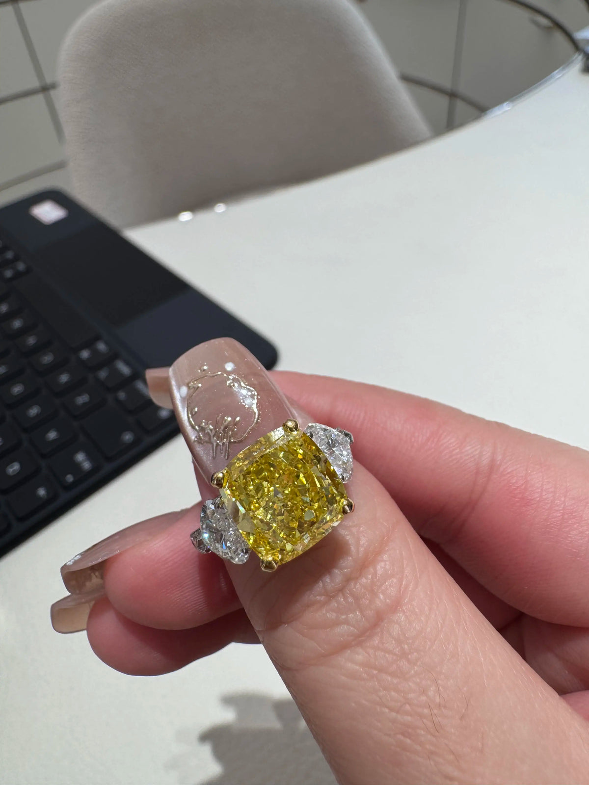 A hand wearing a ring with a large yellow cushion-cut center stone and silver-toned side stones, photographed in an indoor setting with a keyboard and chair in the background.