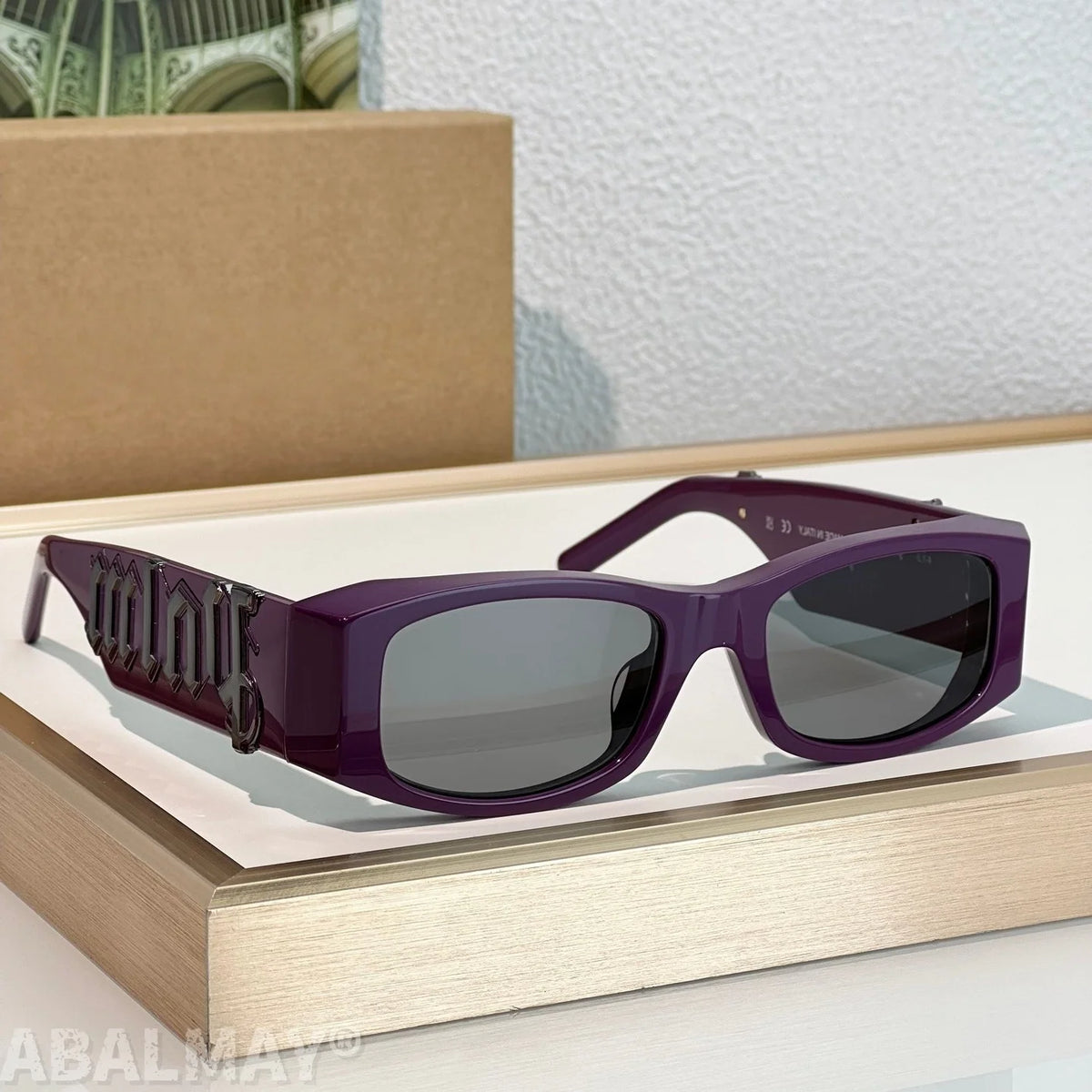 Purple rectangular sunglasses with dark lenses and decorative lettering on the temple arm, displayed on a light wood surface with a cardboard box in the background.