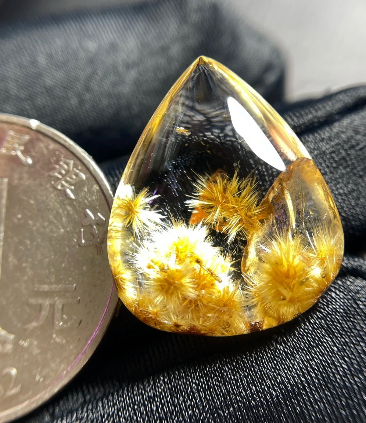 A teardrop-shaped clear gemstone pendant containing yellow dried flower buds and dark inclusions, shown next to a coin for scale on a dark textured fabric background.