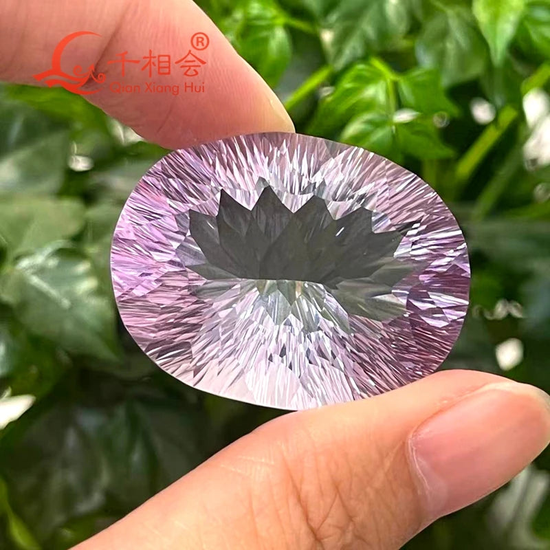A faceted oval gemstone with a pink-lavender outer halo and a dark gray central lotus-like pattern, held between two fingers against a background of green leaves. A red logo for 'Qian Xiang Hui' is visible in the top left corner.