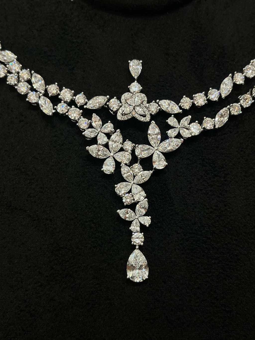 A sparkling silver-tone diamond necklace with a floral and leaf motif, featuring a central teardrop diamond pendant, displayed on a black velvet background.