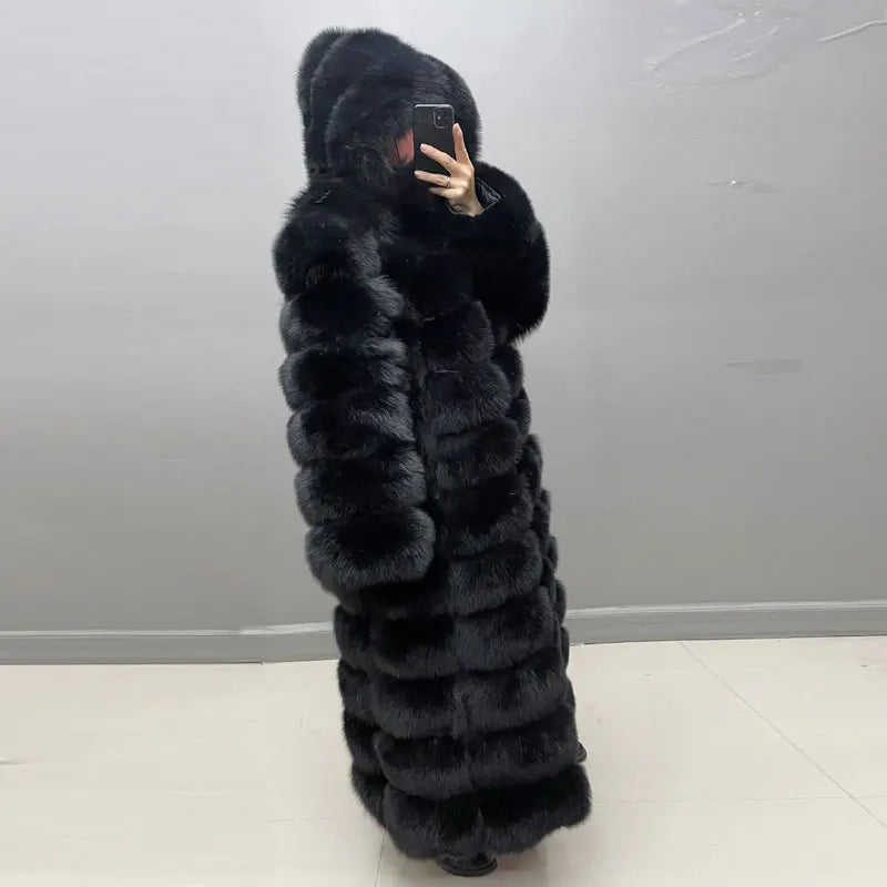 A person wearing a long, black, hooded faux fur coat takes a mirror selfie in an indoor setting with a gray wall and light-colored floor.