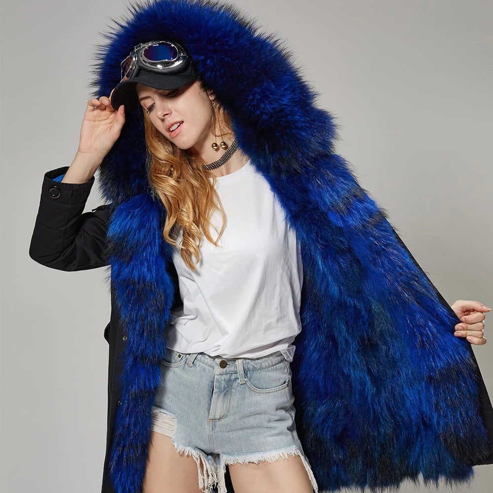 A woman models a luxurious bright blue fur coat with a large matching fur-lined hood, styled with a black coat underneath, a white t-shirt, denim shorts, and goggles on a cap, against a neutral gray studio background.