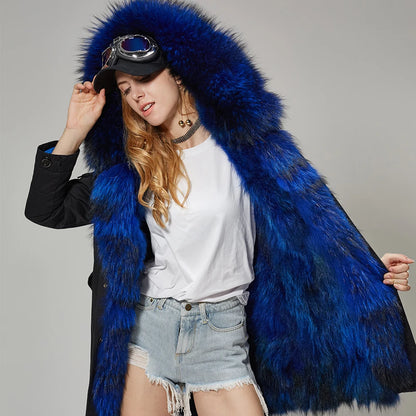 A woman models a luxurious bright blue fur coat with a large matching fur-lined hood, styled with a black coat underneath, a white t-shirt, denim shorts, and goggles on a cap, against a neutral gray studio background.