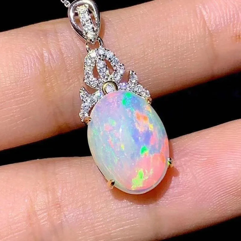 A close-up of a silver pendant necklace with a large, round opal gemstone showing vibrant iridescent colors, held on a person's fingers against a black background.