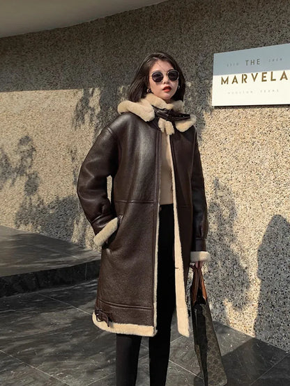 A woman wearing a dark brown shearling-lined leather coat with a beige fur-trimmed collar, standing outdoors against a textured stone wall.
