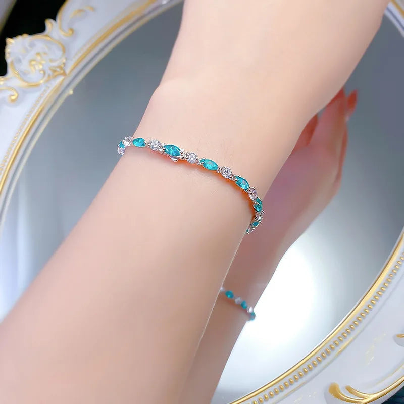 A close-up of a person's wrist wearing a delicate bracelet with marquise-cut turquoise blue stones and small clear accent stones in a silver-toned setting, with a matching bracelet faintly visible in a mirror.