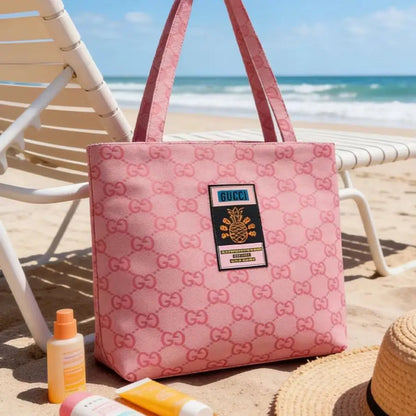 A pink canvas tote bag with an allover GG monogram pattern and a central patch reading 'GUCCI', placed on a sandy beach next to a white lounge chair, sunscreen bottles, and a straw hat.