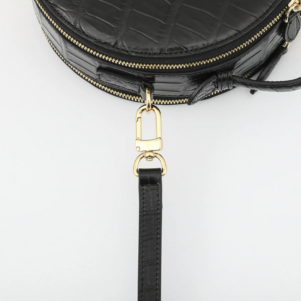 Close-up of a black crocodile-embossed leather handbag with gold-tone hardware and a detachable black leather wristlet strap, shown on a light gray background.