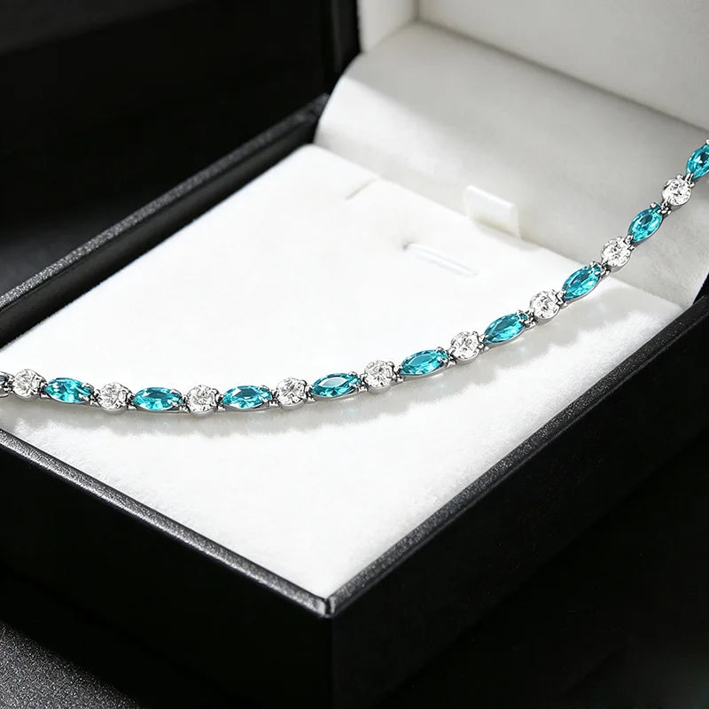 A silver tennis necklace with marquise-cut turquoise blue gemstones and small clear accent stones, displayed inside an open black jewelry box with a white velvet insert.