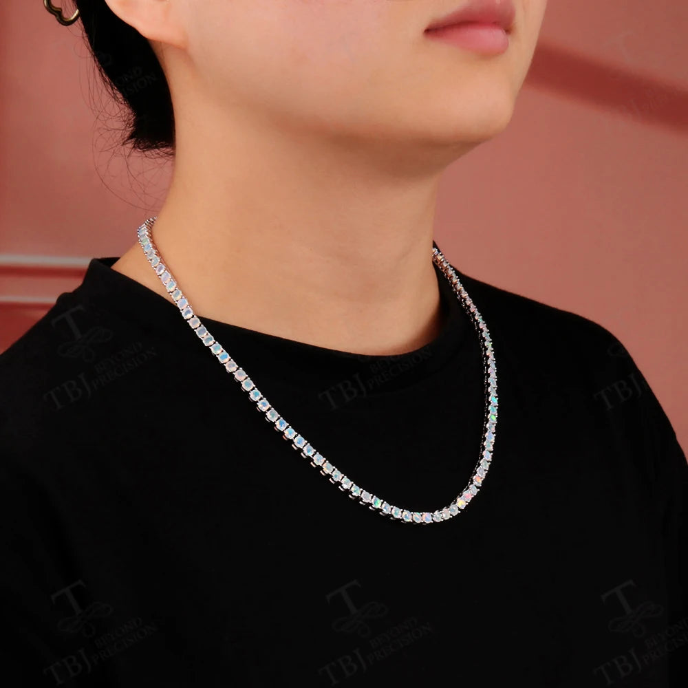 A close-up of a person wearing a silver-toned tennis-style necklace with clear rectangular stones, shown against a black shirt and a soft pink background.