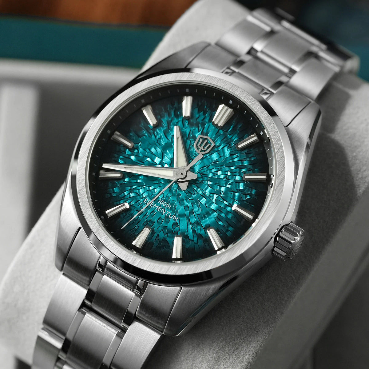 A close-up of a stainless steel wristwatch with a textured teal and turquoise sunburst dial, displayed on a gray cushion.