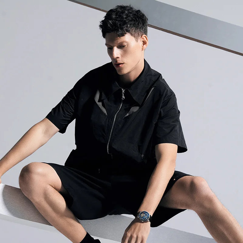 A man wearing a black short-sleeve collared zip-front shirt and matching black shorts, seated on a white platform against a light gray background.