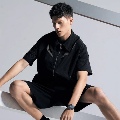 A man wearing a black short-sleeve collared zip-front shirt and matching black shorts, seated on a white platform against a light gray background.