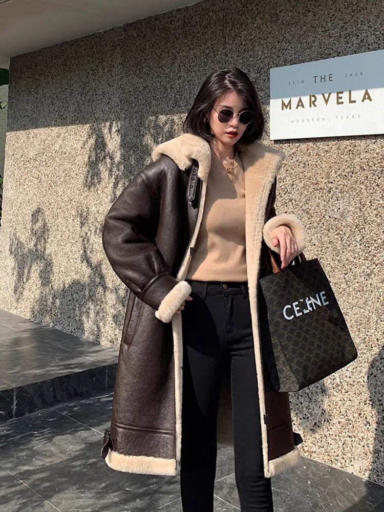 A woman wearing a brown shearling-lined coat with beige fur trim, a beige top, black pants, sunglasses, and holding a black Celine tote bag, standing outdoors in front of a textured wall with a sign that reads 'THE MARVELA'.
