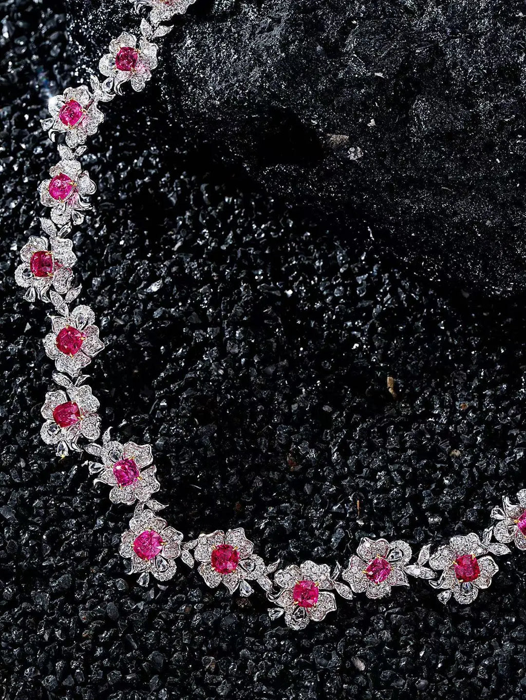 A close-up of a floral necklace with bright pink gemstones and silver-toned settings, arranged on a dark textured surface.