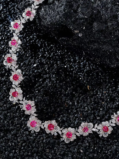 A close-up of a floral necklace with bright pink gemstones and silver-toned settings, arranged on a dark textured surface.