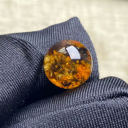 A round resin cabochon featuring dried botanical elements inside, resting on a textured dark gray fabric surface.