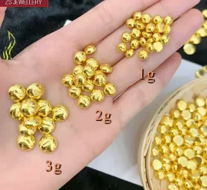 A hand holding three piles of small shiny gold round beads labeled 1g, 2g, and 3g, with a basket of more gold beads at the bottom right. A small 'ZS JEWELLERY' label appears in the top left.