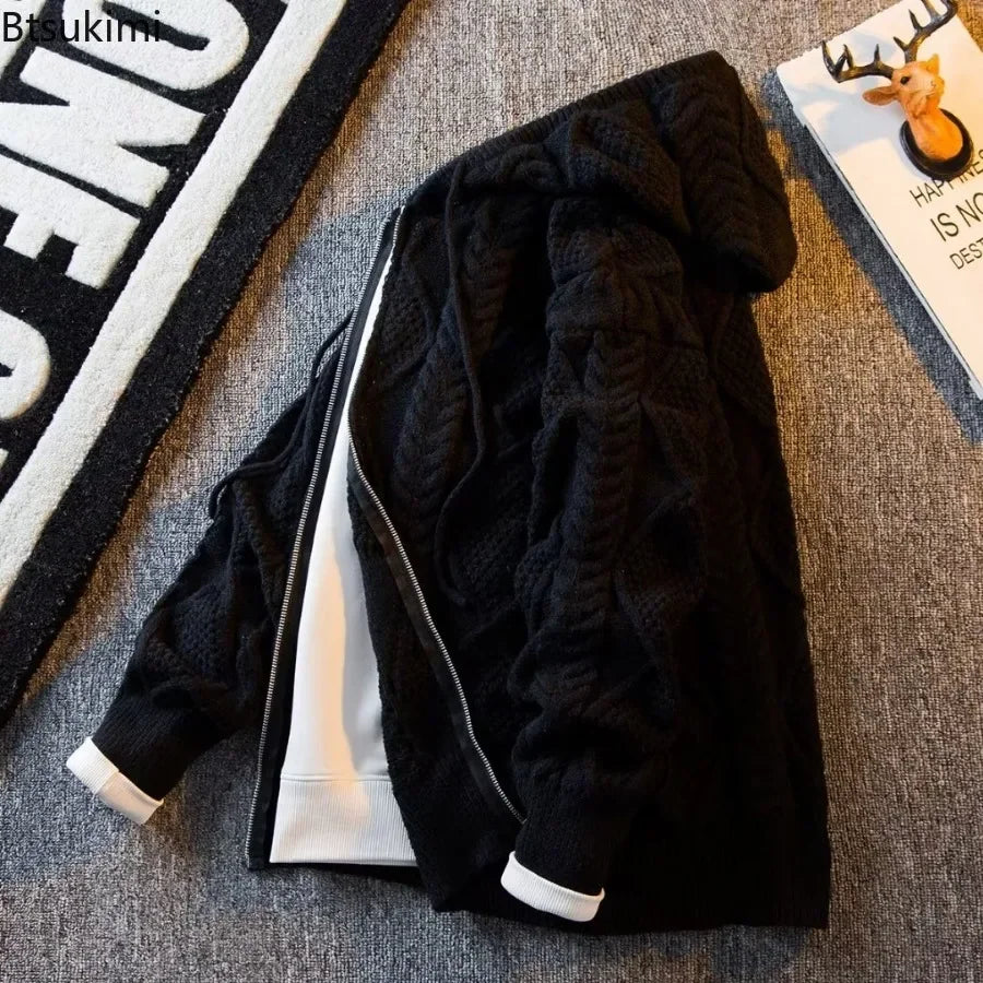 A black cable-knit zip-up hoodie jacket with a white inner lining and white cuff trims, laid flat on a gray textured carpet next to a rug and a magazine.