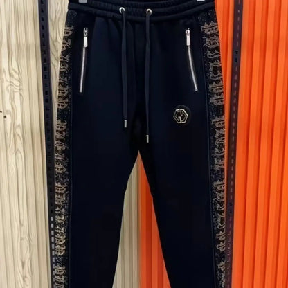 Black jogger sweatpants with decorative side panels featuring a repeating beige monogram pattern, dual silver zippers, and a small hexagonal logo patch on the left thigh.