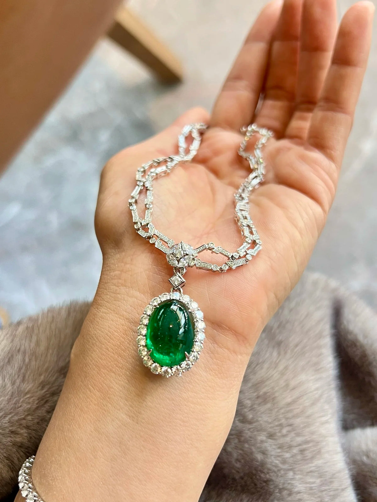 A close-up of a hand holding a silver-toned chain necklace with a large oval green gemstone pendant surrounded by small clear stones.