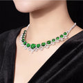 A close-up of a model's neck wearing a silver-toned choker necklace with large green emerald-cut gemstones and small clear accent stones, paired with a black velvet top against a dark background.