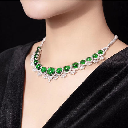 A close-up of a model's neck wearing a silver-toned choker necklace with large green emerald-cut gemstones and small clear accent stones, paired with a black velvet top against a dark background.