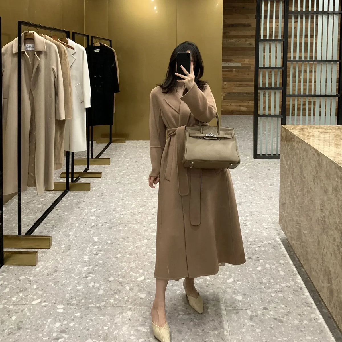 A woman takes a mirror selfie in a boutique wearing a long taupe-brown wrap coat dress and carrying a matching structured handbag. The boutique interior features neutral beige and brown tones, with clothing racks visible in the background.