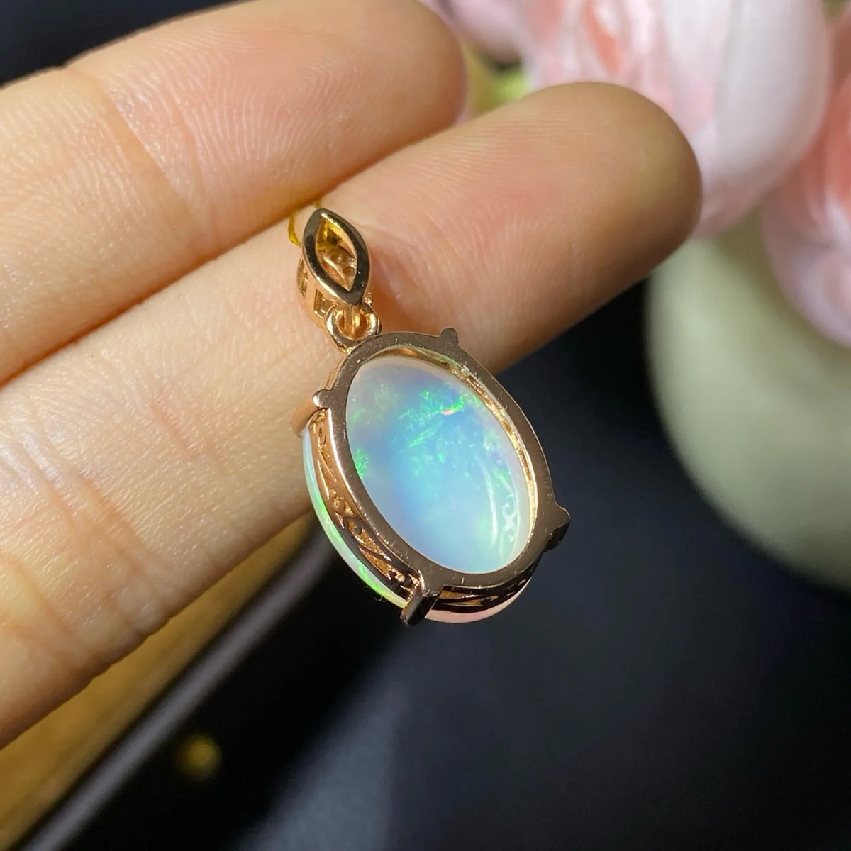 A rose gold pendant with an oval iridescent opal gemstone, held between fingers against a dark, blurred background with soft pink flowers.
