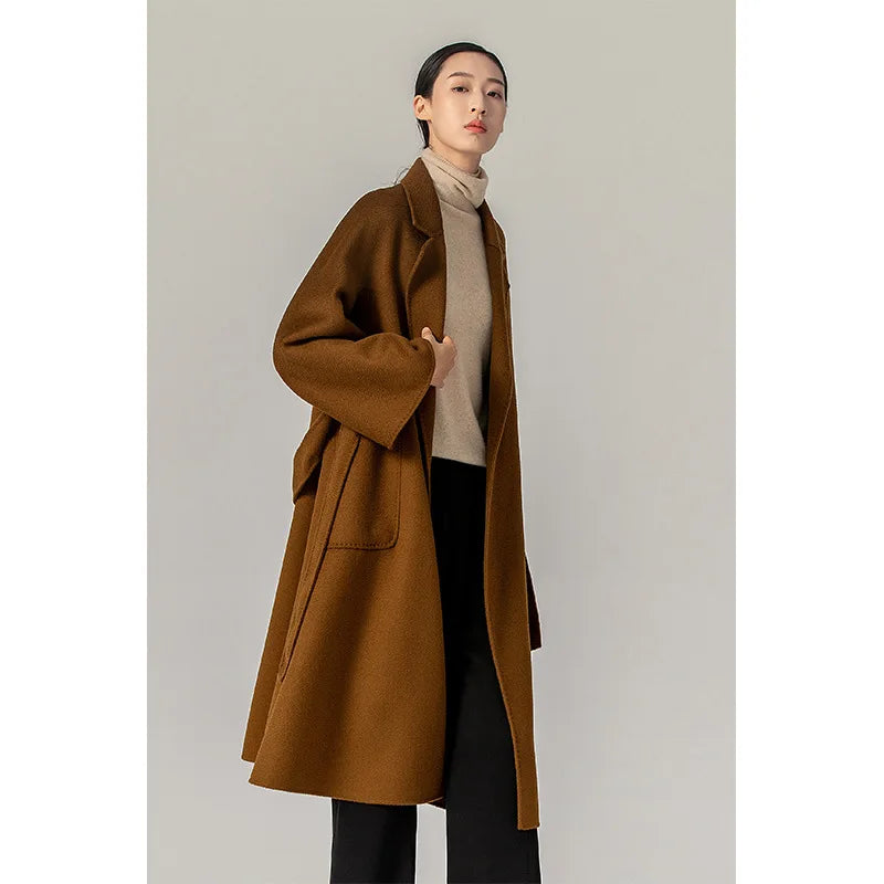 A woman modeling a long brown wool-blend coat over a beige turtleneck sweater and black pants, standing against a plain light gray studio background.