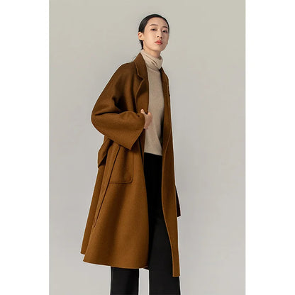 A woman modeling a long brown wool-blend coat over a beige turtleneck sweater and black pants, standing against a plain light gray studio background.