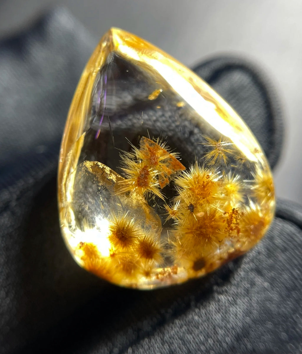 A teardrop-shaped clear resin gemstone containing golden-yellow dried flower spires, resting on a black textured surface.