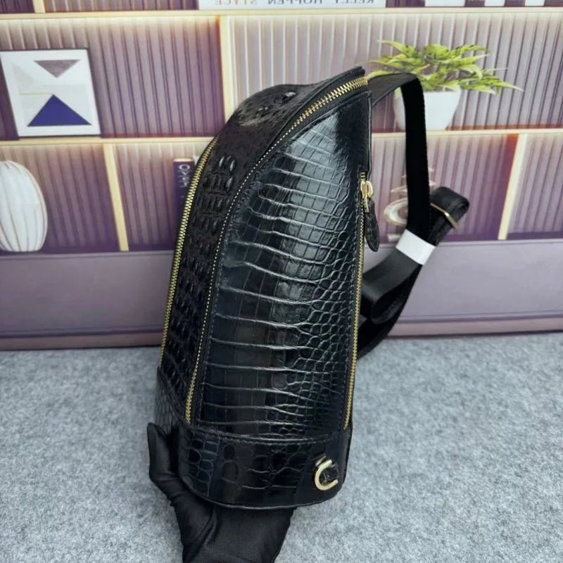 A black crocodile-embossed leather sling backpack with gold zippers, shown with a black strap and a small gold C-ring detail, set against a purple paneled background.