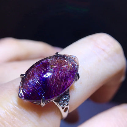 A close-up of a silver ring with a large, marquise-shaped purple iridescent gemstone held on a finger against a dark background.