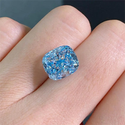A loose cushion-cut light blue gemstone displayed on a person's finger against a dark gray background.