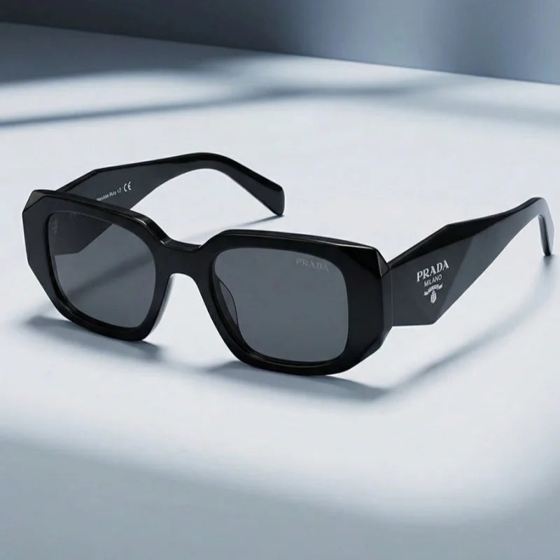 Prada sunglasses with a glossy black acetate frame, dark gray lenses, and the Prada logo printed on the temple arm, displayed on a clean light gray surface.