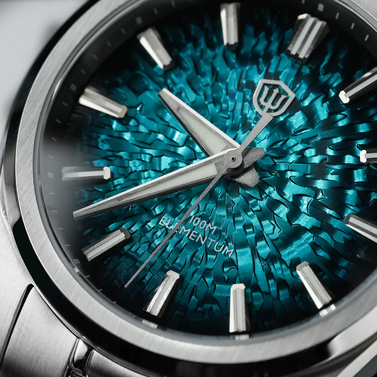 Close-up of a wristwatch dial with a metallic silver-toned case, silver hour markers and hands, and a vivid blue-green textured cracked-glass style face. The dial shows the brand mark and the text '100M ELEMENTUM'.