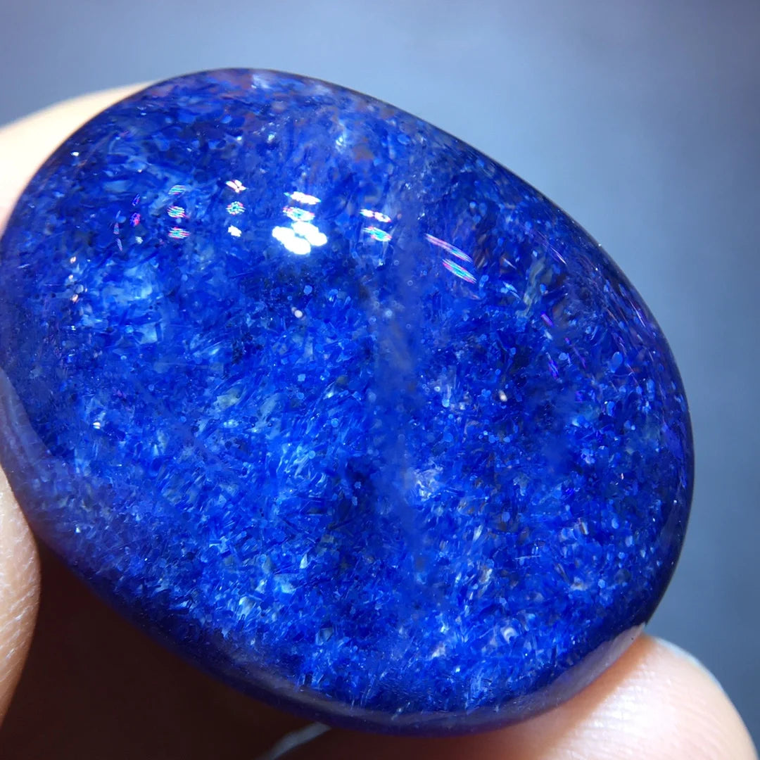 A polished oval-shaped blue stone held between fingers, showing a highly textured, crystalline surface with bright blue and lighter icy blue highlights under direct light.