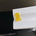 A rectangular yellow gemstone with a rough cushion cut, showing vibrant yellow color and sparkling facets, displayed on a white textured surface.