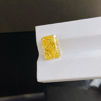 A rectangular yellow gemstone with a rough cushion cut, showing vibrant yellow color and sparkling facets, displayed on a white textured surface.