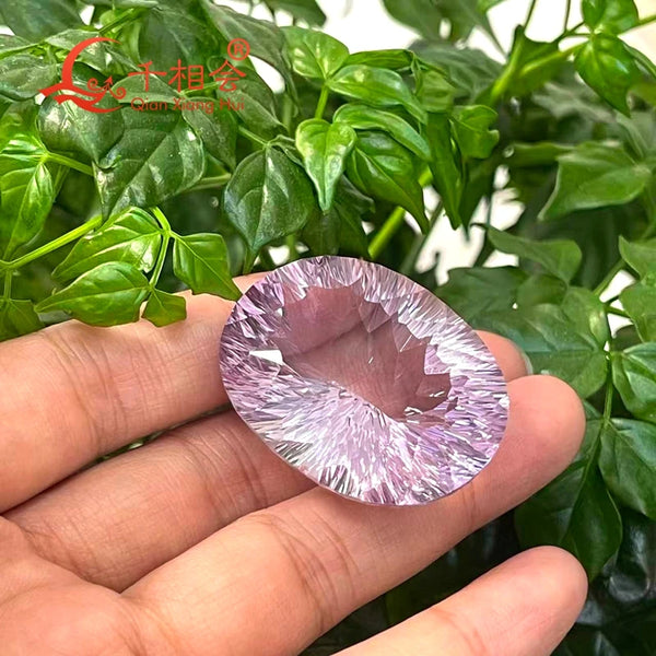 A faceted oval gemstone with a lavender-purple center and radiating clear crystal points, held in a person's hand against a background of green leaves. A red and gold 'Shan Yang Hu' logo is visible in the top left corner.