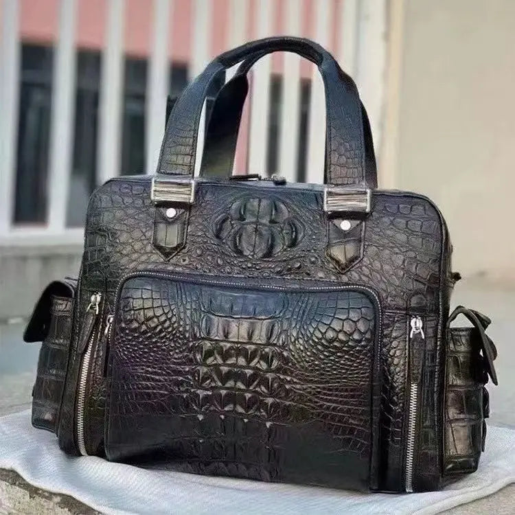 A black crocodile-embossed leather briefcase-style handbag with dual top handles, silver-tone hardware, and multiple zippered pockets, photographed outdoors on a light surface with a blurred building background.