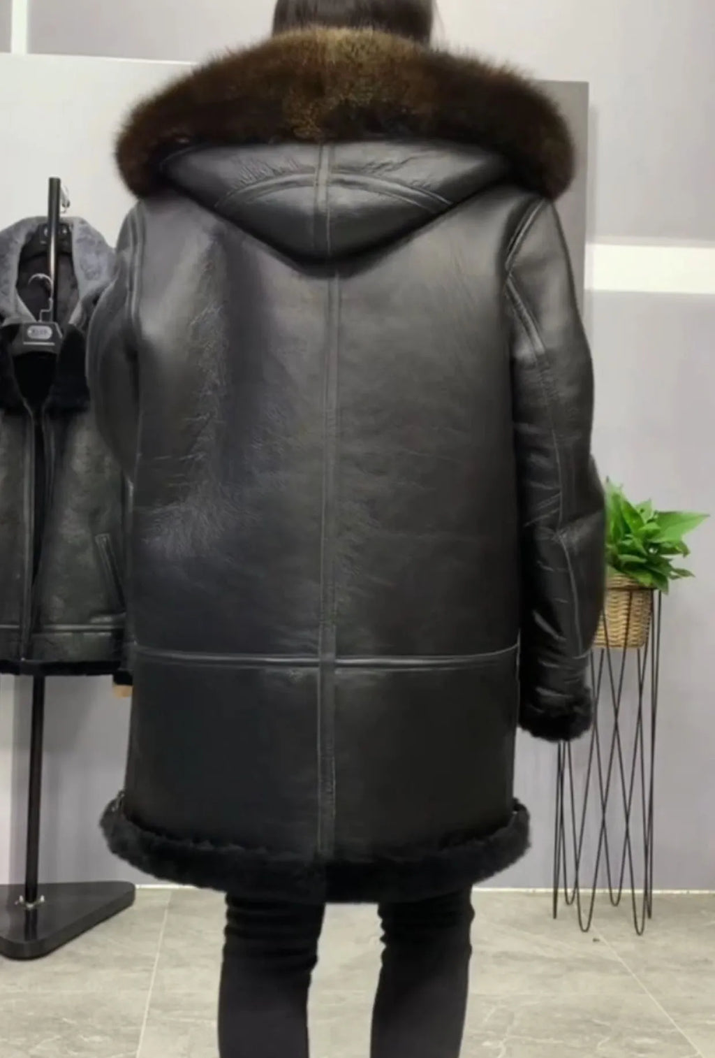 A person shown from the back wearing a black leather hooded coat with a brown fur-trimmed hood and hem, standing indoors in a store-like setting.