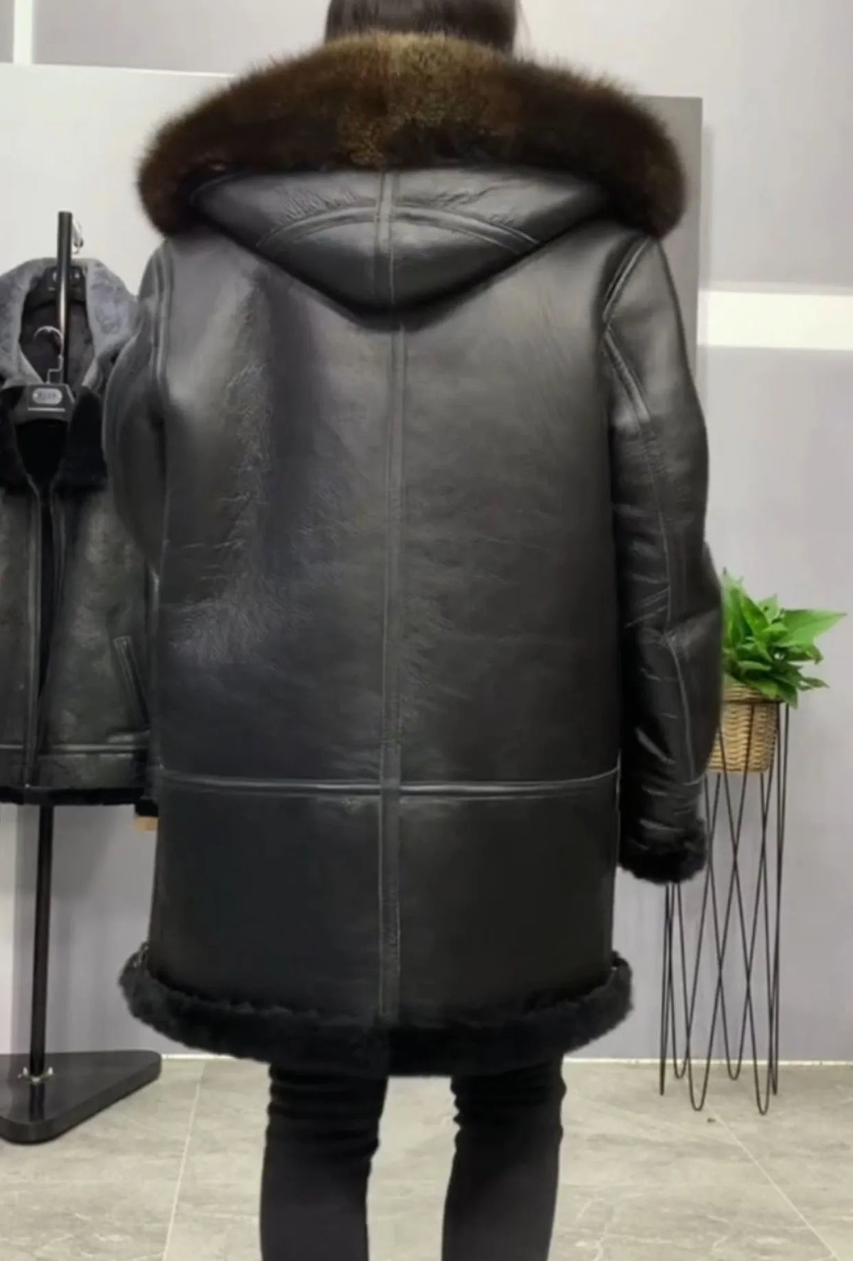 A person shown from the back wearing a black leather hooded coat with a brown fur-trimmed hood and hem, standing indoors in a store-like setting.