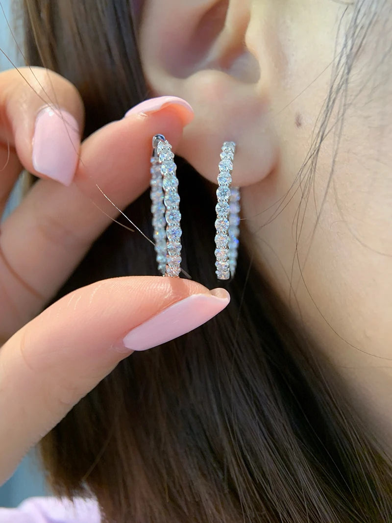 A close-up lifestyle shot of a person wearing silver hoop earrings lined with clear iridescent rhinestones, with a hand holding the earrings to show them more clearly.