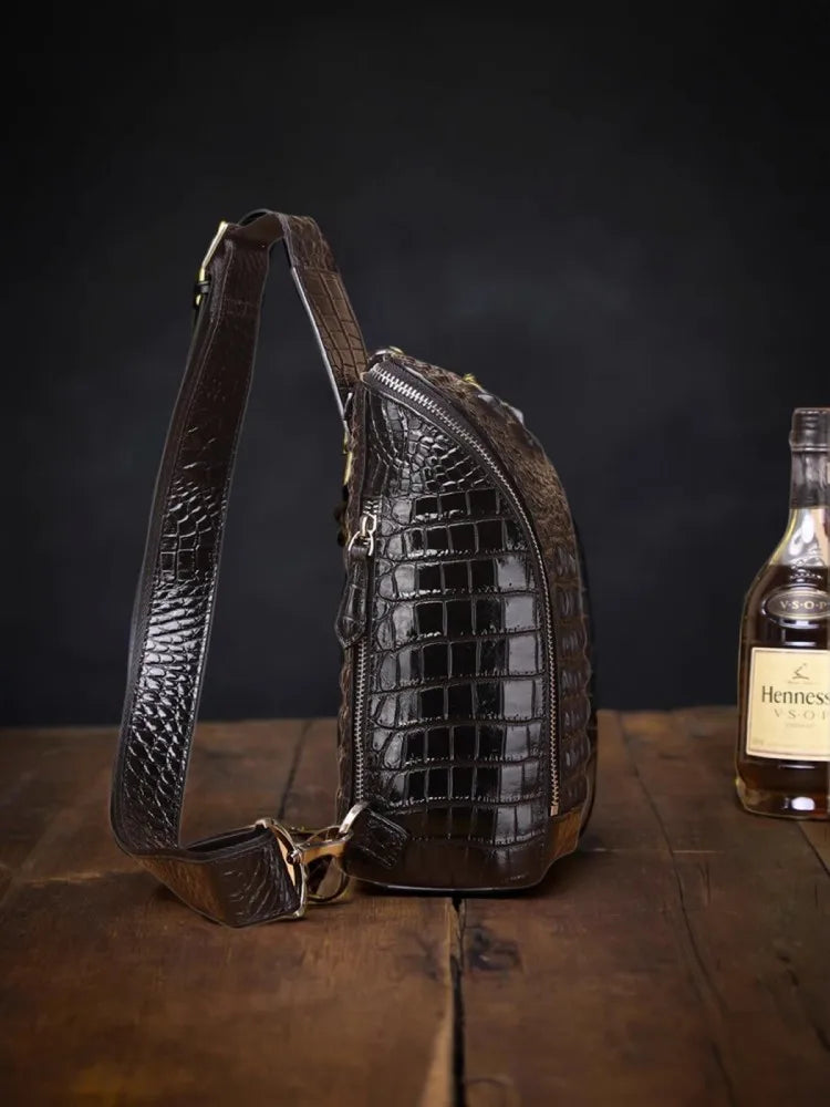A dark brown crocodile-embossed leather sling bag with a matching shoulder strap, displayed on a rustic wooden table next to a bottle of Hennessy cognac.